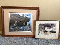 Two framed art prints leaning against a beige wall on a carpeted floor, showing the larger pelican and ship image on the left and smaller boat print on the right. Both have wooden frames; the larger frame is golden-brown, the smaller frame is gold-colored.