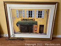 Framed artwork showing a yellow building facade with blue shutters, green awning, and 'Le Village' sign, resting on floor against wall.