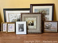 Photo of seven framed artworks of varying sizes displayed on a wooden floor against a yellow wall, showing floral and nature themes with various frame styles.