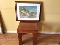 Framed coastal artwork displayed on wooden table against yellow wall, showing framing, matting and overall condition.