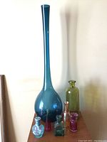 Overall view showing seven glass vases and bottles of different sizes and colors clustered together against a wall.