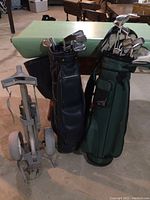 Two golf bags standing upright filled with golf clubs and a gray golf trolley next to them on a concrete surface.