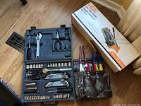 Photo showing opened black plastic tool case containing assorted hand tools and part of HDX tile cutter box in background on wooden floor.