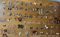 Photo shows 50+ pairs of assorted earrings spread on wooden surface with studs, clips, hoops and dangles in used condition.