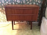 Overall front view of the mid-century wooden side table showing two drawers and tapered legs, background floral wallpaper