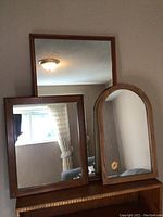 Three wooden framed mirrors placed on a wooden surface against a beige wall. Large rectangular mirror at the back, smaller rectangular mirror and an arched-top mirror in front.