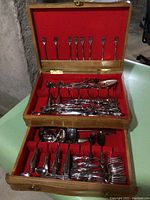 Overall view of open wooden case containing various plated stainless steel flatware including forks, spoons, and serving pieces, organized in compartments with red lining.