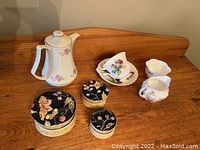 A collection of assorted ceramic items including a white floral teapot, three floral trinket boxes with black lids, and a cream and sugar set with delicate flowers.
