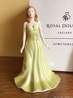 Front angled view of Royal Doulton porcelain figurine in pale green-yellow gown, with green gemstone on bodice, showing detail and box in background.