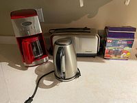 Photo showing red drip coffee maker, stainless steel electric kettle, toaster, and boxed sandwich maker on countertop.