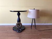 Side view of round black wooden side table with pedestal base, tripod floor lamp with beige shade, decorative mirror and decorative bowl on floor, showing dust and scratches on table