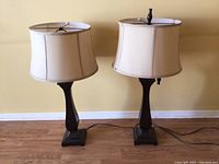 Pair of two used table lamps with beige fabric shades and dark wood bases, sitting on wooden floor in front of yellow wall.