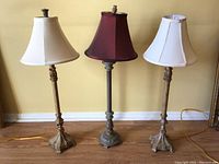 Three table lamps shown together, metal bases with classical design, two white shades, one dark red shade.