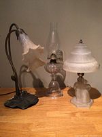 Three table lamps shown together. Left: electric lamp with tulip-shaped cream glass shade and dark metal stand. Middle: clear glass oil lamp with chimney and metal burner. Right: frosted glass electric lamp with embossed glass shade and figurine base.