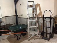 Photo showing side view of the green metal wheelbarrow with wooden handles, aluminum ladder, and black hand truck dolly.