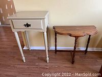 Two wooden tables displayed side by side, one beige with drawer and one natural wood with curved top edge, on a hardwood floor against a plain wall