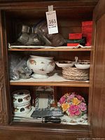 Full view of wood cabinet shelves showing Royal Albert China pieces including a pedestal bowl, small bowls, stacked trivets in a wicker basket, metal pet bowls, place card holders, picture frame, cutlery sharpeners, and a floral ceramic trivet.