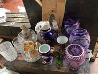 Overview of lot showing a variety of crystal vases and bowls, colored glass, and a blue China vase with gold decoration and broken top.