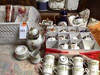 Overview of assorted china and glassware items including tea sets, creamers, sugar bowls, and crystal pieces in protective cases and on carpeted floor.