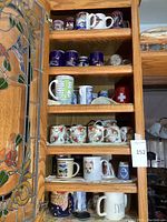 Multiple shelves showing a variety of coffee mugs including floral, scenic, and novelty designs, arranged in a cabinet.