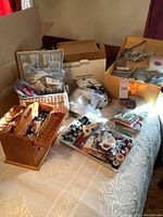 Overview photo of sewing supplies on bed including boxes, wooden sewing box, plastic trays and bags of sewing notions.
