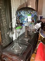 Photo showing the stained glass table lamp with churches and foliage design on the shade, beside two twin solar lamps with clear ribbed glass shades and metal frames.