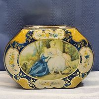 Front view of antique hinged lid candy tin with painted artwork depicting two women and floral motifs.