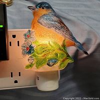 Front view of the glowing bird-shaped glass night light plugged into an electrical outlet, showing detailed colors and textured glass.