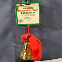 Close-up photo showing the brass bell with red bow hanging, including the original green and white hang tag with product information and usage instructions for indoor or outdoor use.