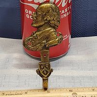Front angle of brass wall hook showing profile bust of English gentleman and decorative shield below, mount holes visible.