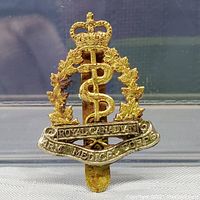 Front view of Canadian Royal Canadian Army Medical Corps cap badge pin, showing gold crown, wreath of maple leaves, and rod of Asclepius symbol with banner inscription.