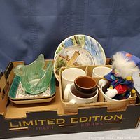 Box holding a green glass swan figurine, square dishes, and a small clown doll with colorful clothes and feathered hat, along with a brown vase and decorative plates behind.