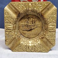 Top view showing square brass-toned ashtray with paddlewheel steamboat relief and decorative edges.