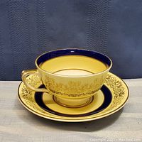 Front view of the porcelain tea cup on its matching saucer showing the cream base, gold floral design, and cobalt blue accents.