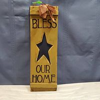 Front view of wood sign painted yellow with 'BLESS OUR HOME' text, star cutout, and checked bow