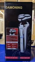 Front of box showing the Damonning electric shaver DMN-5800 with its 4D omni-directional intelligent floating shaving system. The box mentions USB and DC charging adapters and that the device is 100% body washable.