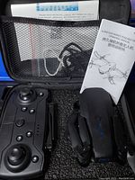 Foldable drone with black remote controller, folded drone body, USB charger cable, and user manuals visible inside foam case