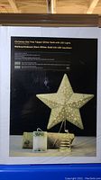 Box image showing the gold star tree topper with LED lights and spring base. Text on box identifies item as Christmas Star Tree Topper Glitter Gold with LED Lights.