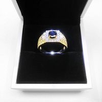 Front angle of gold plated men's ring size 10 showing blue center stone and clear stones around it in black velvet cushion and white box.