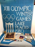 Full view of the Olympic Winter Games 1980 blue and white woven wall hanging with pins, magazines, a glass, and other memorabilia displayed in front