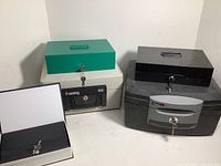 Photo showing five safes and boxes including green and black metal boxes, a beige Sentry model 1150 safe, a gray fire box, and a black book safe.