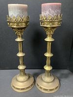 Pair of tall brass candle holders with pillar candles placed on top against a dark background, showing overall look and height.