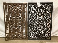 Photo showing both cast iron entry mats leaning vertically, displaying full decorative patterns and size comparison.