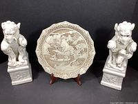 Shows pair of white ceramic Foo dog bookends and Ivory Dynasty decorative plate on wooden stand against black background.