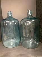 Pair of large 5 gallon glass bottles shown from front, clear with embossed branding visible.