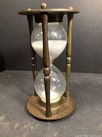 Front view of brass and glass hourglass showing white sand inside glass bulbs held by brass frame with four vertical supports.
