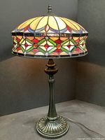 Front view of the lit stained glass table lamp showing colorful pattern on the shade and decorative metal base.
