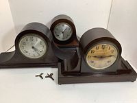 Three mantle clocks on a white surface with two winding keys placed in front.