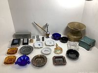 Wide shot showing full collection arranged on white surface, including brass spittoon, pipe, ashtrays and boxes with lighter cases.