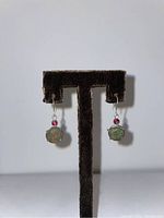 Front view of pair of earrings displayed on brown textured stand, showing antique coins with green patina and small red bead above each.